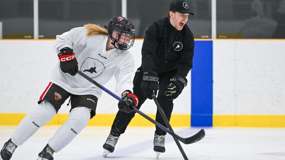 A Perfect Skating hockey player and their coach on an indoor ice rink, one in a white jersey and black pants, the other in a black jersey and pants.