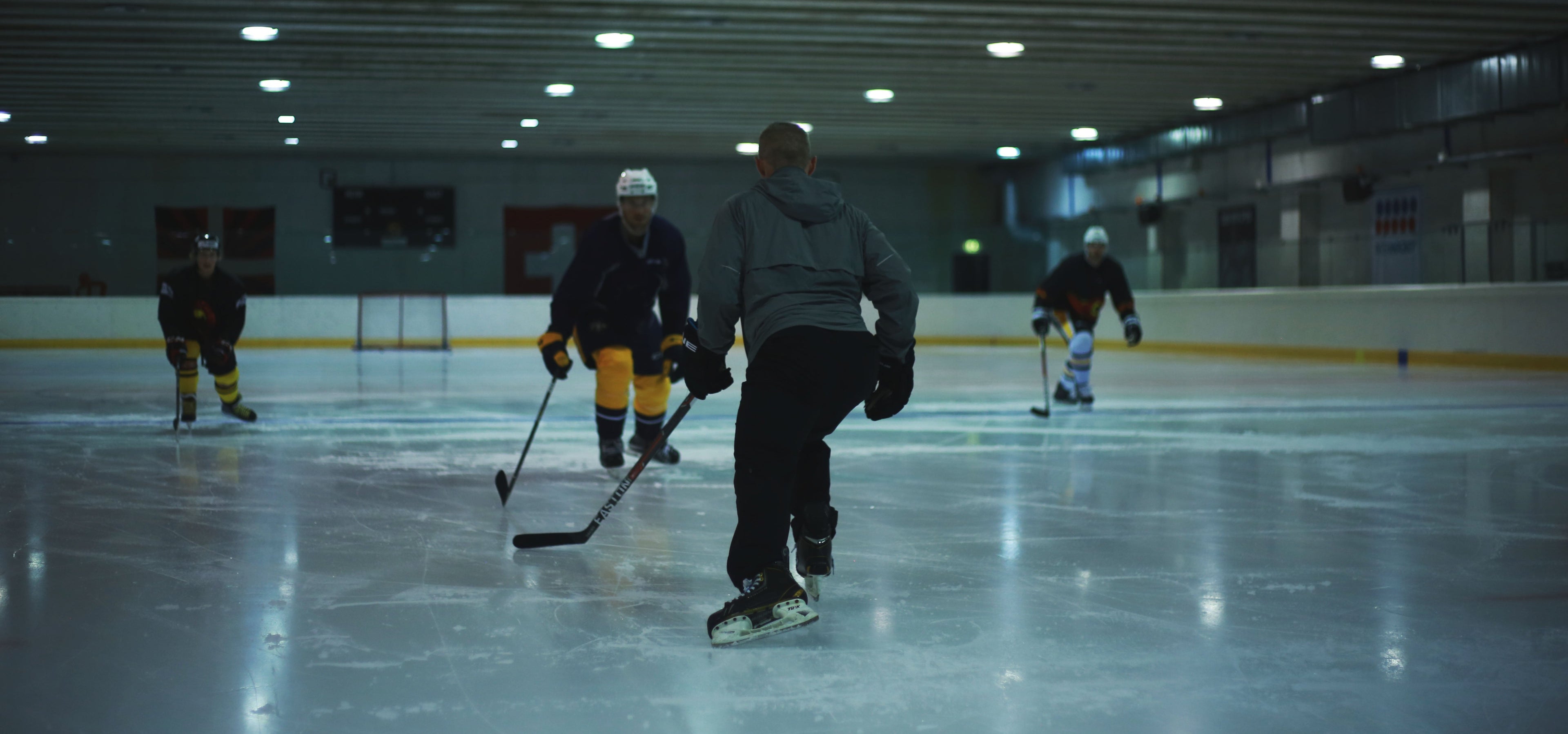 Shawn Allard on an indoor ice rink holding a hockey stick, instructing NHL hockey players visible in the background under overhead lights.
